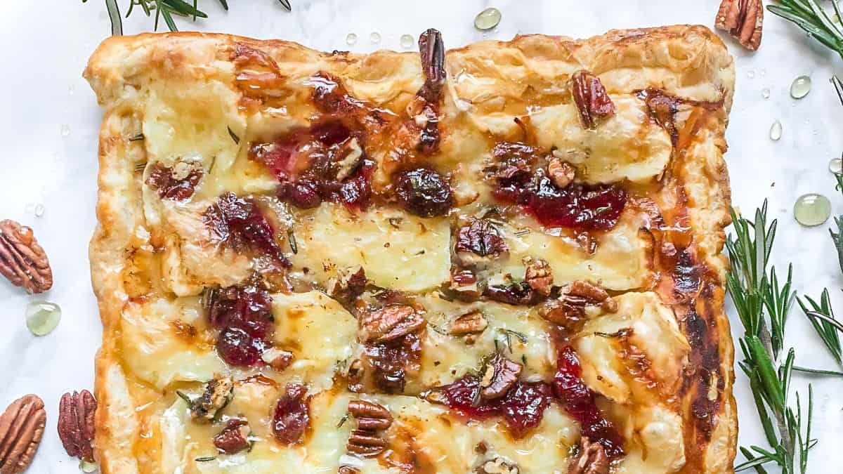 A rectangular puff pastry topped with melted cheese, pecans, and cranberry sauce. Rosemary sprigs and pecans are scattered around it on a white surface.