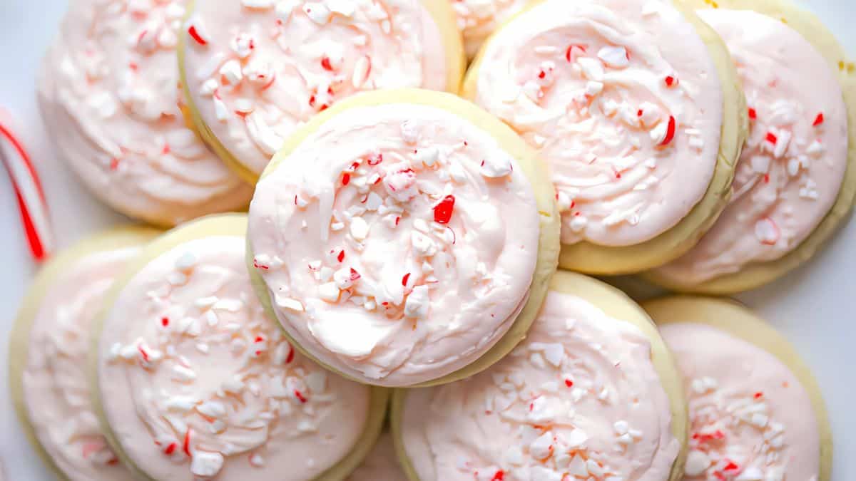 A pile of frosted cookies topped with crushed peppermint candy pieces. The cookies have a soft, inviting appearance and a light pink frosting, creating a festive and sweet treat.