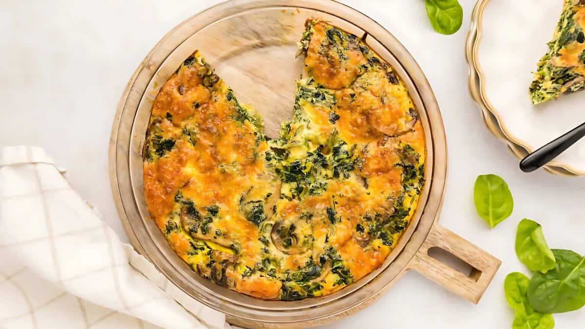 A baked spinach quiche with a slice removed, showing its golden-brown crust and green spinach filling. It's placed on a wooden board, with fresh spinach leaves and a white napkin nearby.