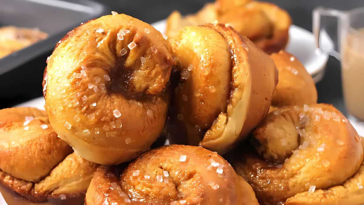 Close-up of a stack of golden, caramel-glazed sticky buns sprinkled with coarse sugar crystals. The pastries have a glossy finish and are piled on a white plate, with a blurred background showing additional buns.