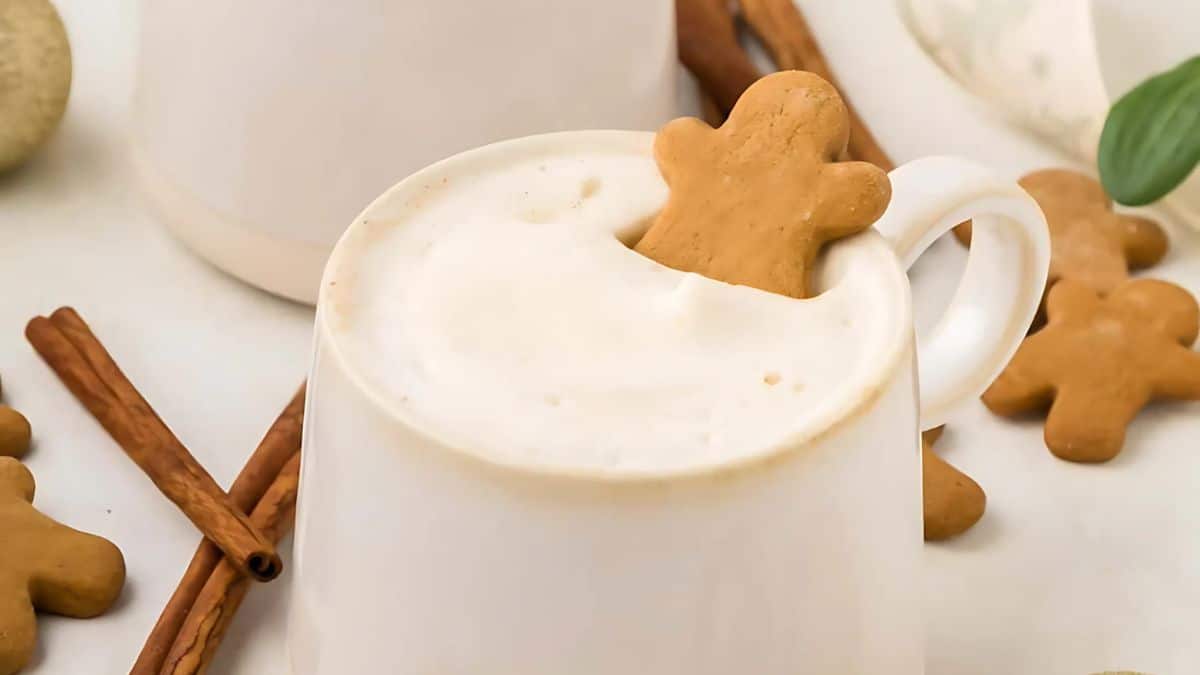A white mug filled with frothy milk, featuring a gingerbread man cookie partially submerged at the edge. Cinnamon sticks are placed beside the mug on a light surface, creating a cozy and festive atmosphere.