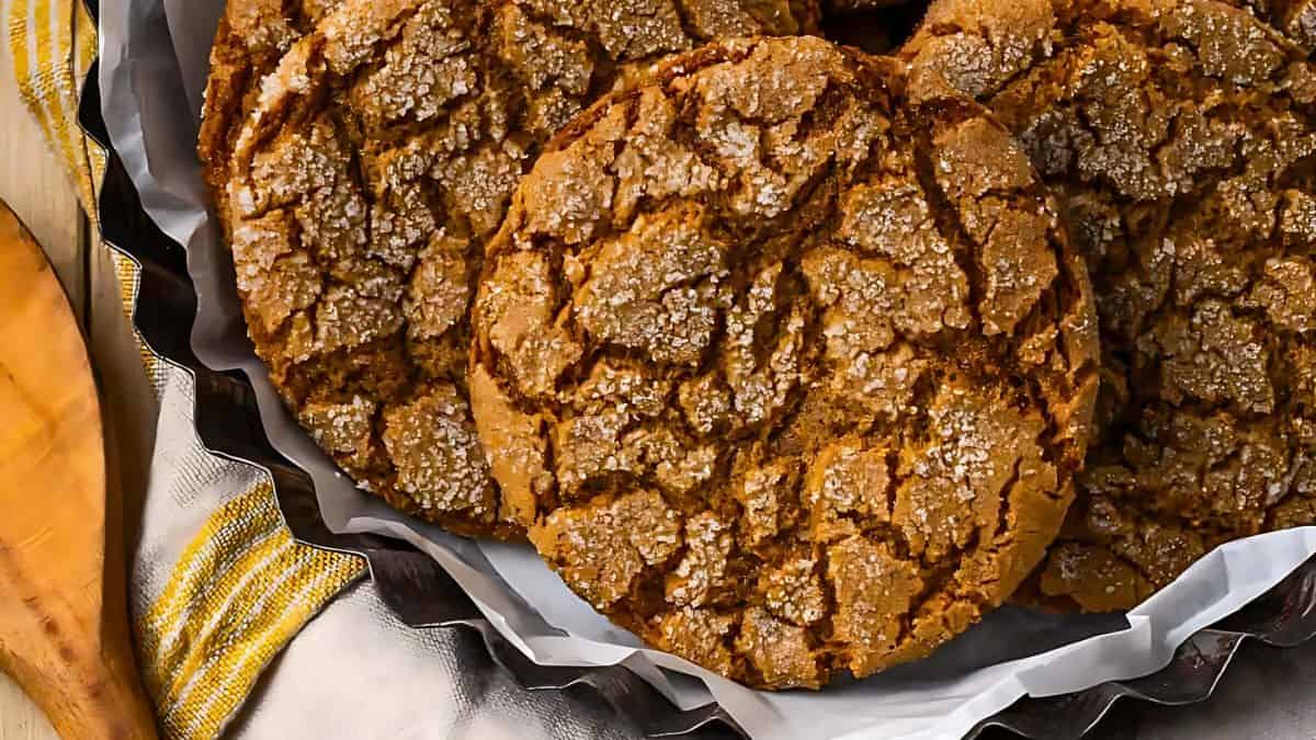 A plate filled with crackled molasses cookies topped with coarse sugar, showcasing a rustic and textured surface. The cookies are surrounded by a decorative cloth and a wooden spatula is partially visible in the background.