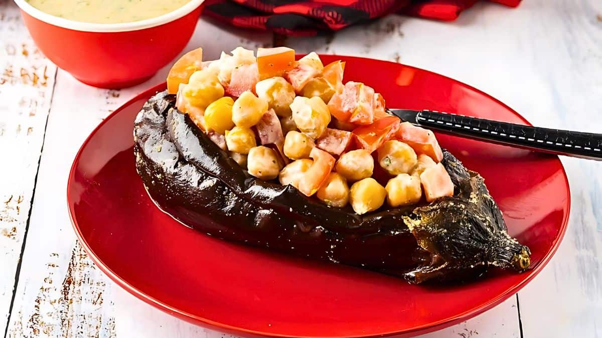 A red plate with a roasted eggplant stuffed with chickpeas and diced tomatoes. A black fork rests on the plate. A red bowl with a creamy sauce is visible in the background on a white wooden table.