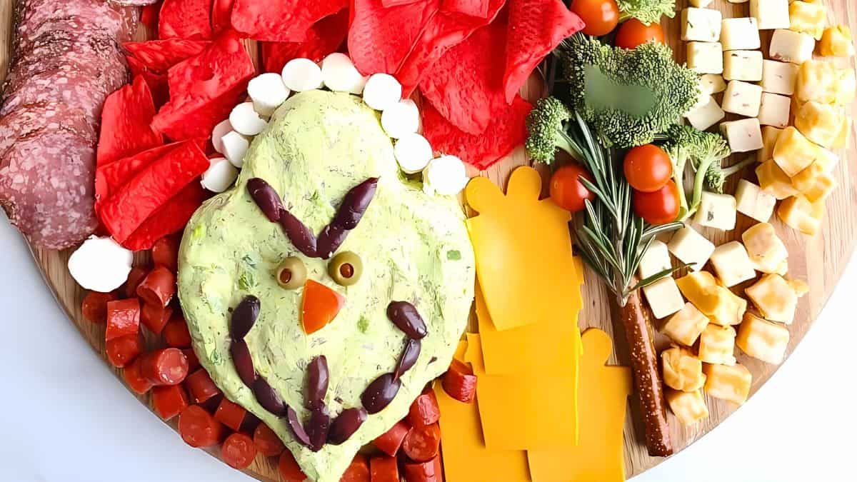 A festive platter features a turkey-shaped dip with olive eyes and pepper beak, surrounded by assorted cheeses, crackers, salami, broccoli, cherry tomatoes, and pepper slices. The vibrant colors create a cheerful holiday presentation.