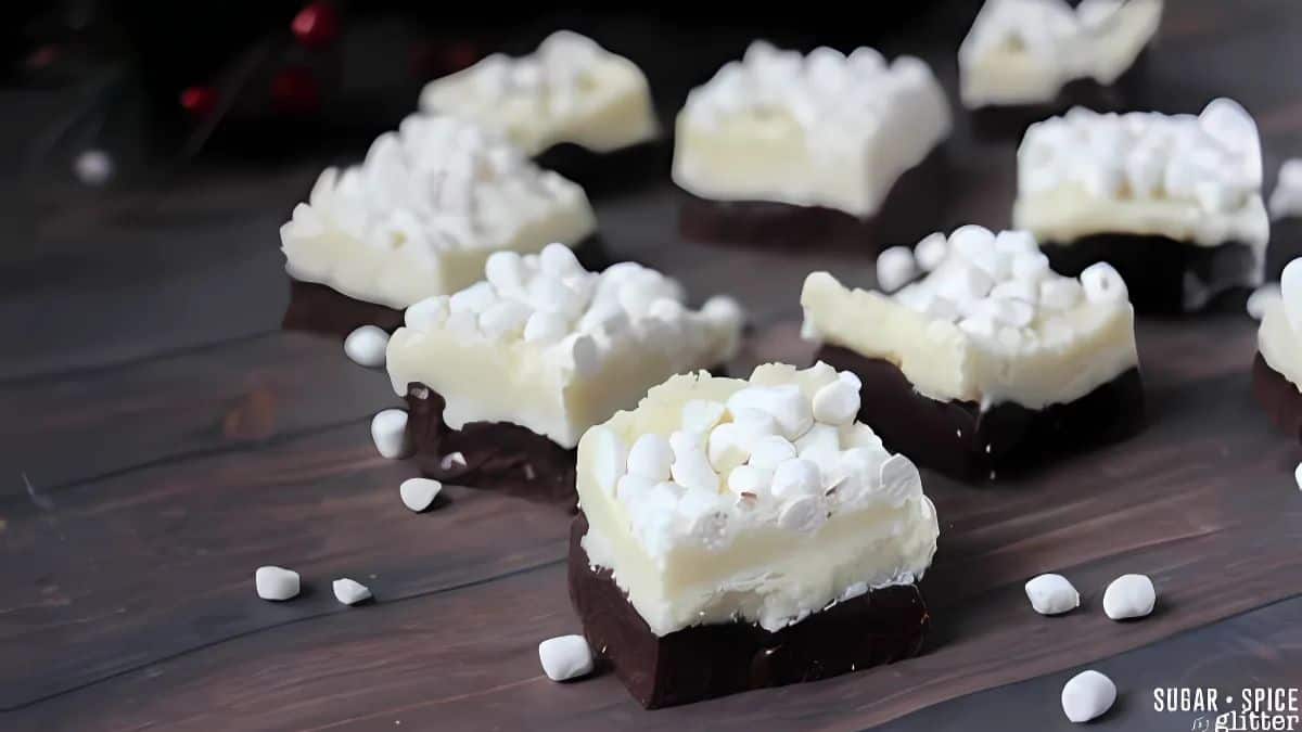 Close-up of white chocolate and dark chocolate layered squares topped with mini marshmallows, scattered on a dark surface. Some marshmallows are spread around, adding to the cozy, homemade treat vibe.