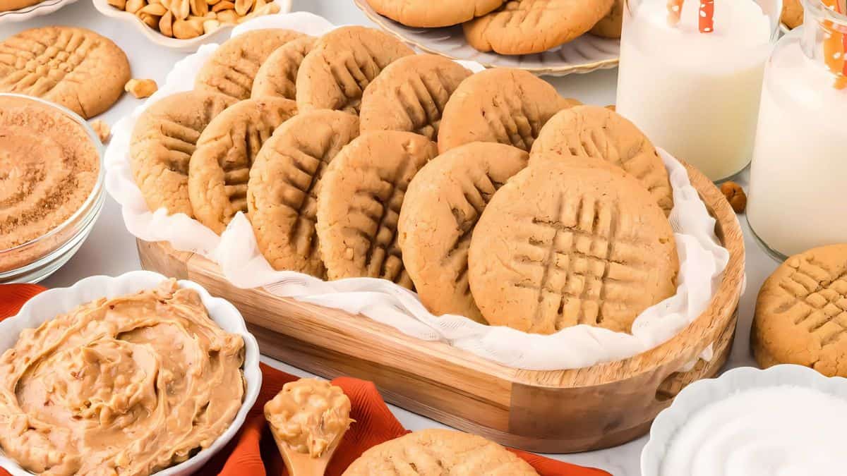 A tray of homemade peanut butter cookies with crisscross patterns is surrounded by a bowl of creamy peanut butter, peanuts, and two glasses of milk with striped straws. The setting is cozy and inviting, suggesting a delicious snack.