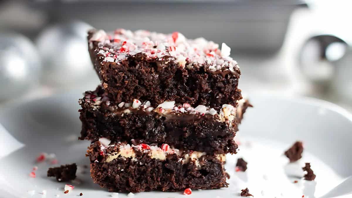 A close-up of three stacked chocolate brownies on a white plate, topped with crushed candy canes. The brownies have a fudgy texture and the candy cane pieces add a festive touch. Silver decorations are blurred in the background.