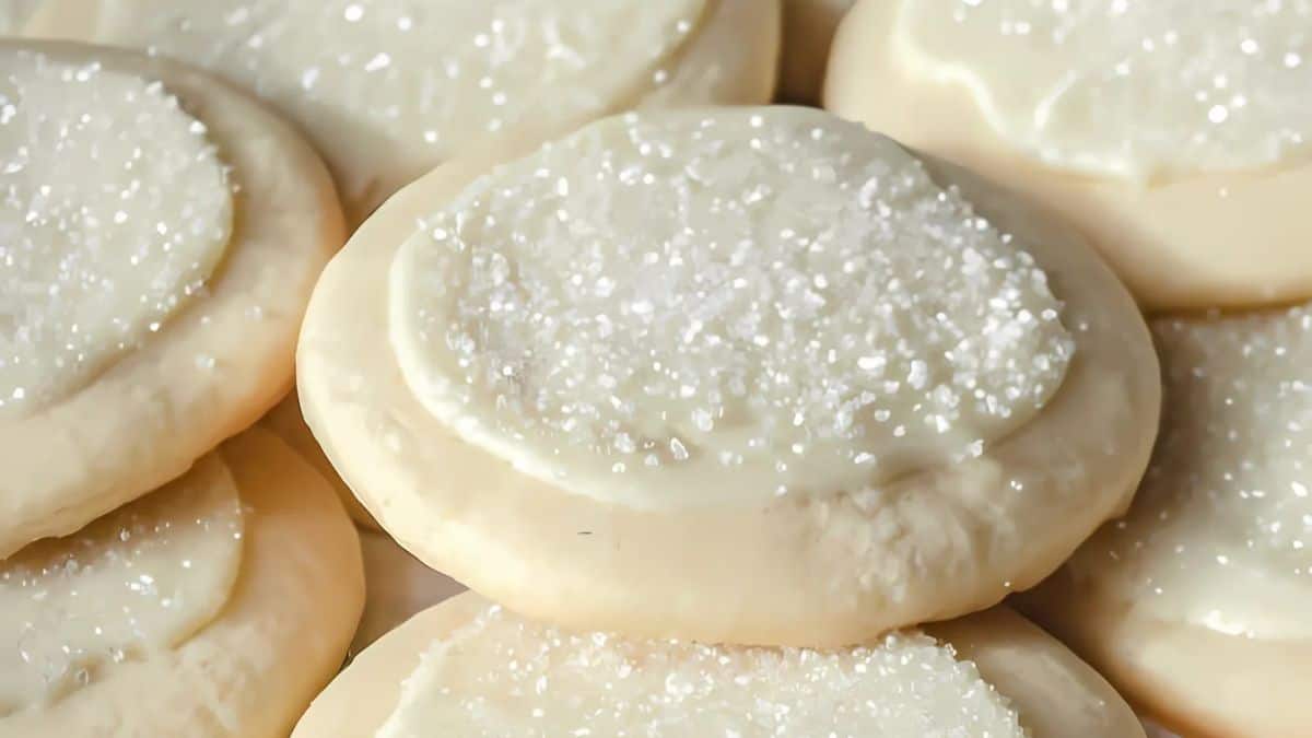 Close-up of several vanilla frosted sugar cookies topped with sparkling white sugar crystals. The cookies appear soft and thick, with a smooth icing layer covering each one.