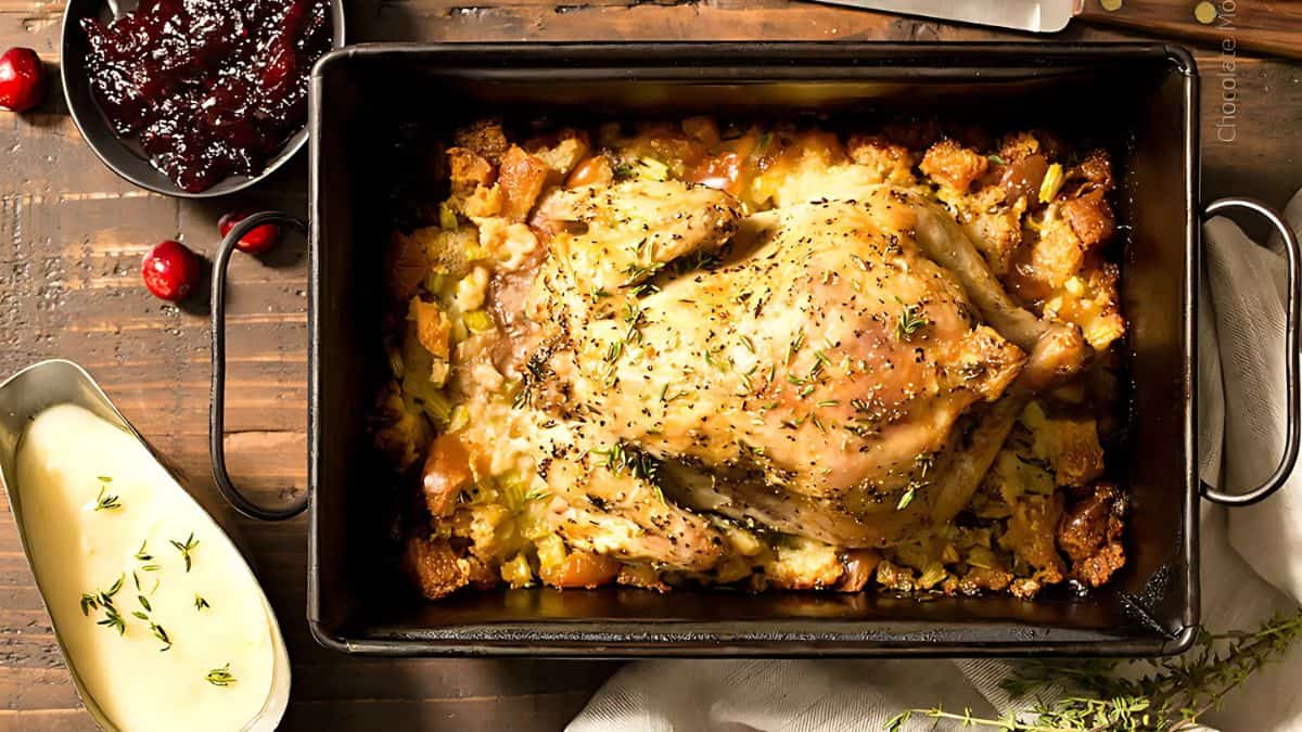 Roast chicken in a baking dish with stuffing, surrounded by a small bowl of cranberry sauce and a gravy boat on a wooden table. Garnished with fresh thyme.