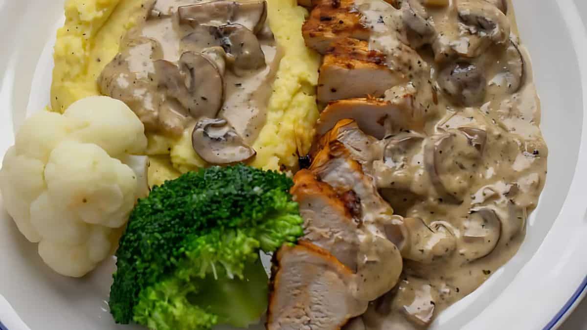 A plate featuring grilled chicken with creamy mushroom sauce, accompanied by mashed potatoes, broccoli, and cauliflower. The chicken is sliced and covered in the rich, herb-infused sauce, complementing the vibrant green vegetables.