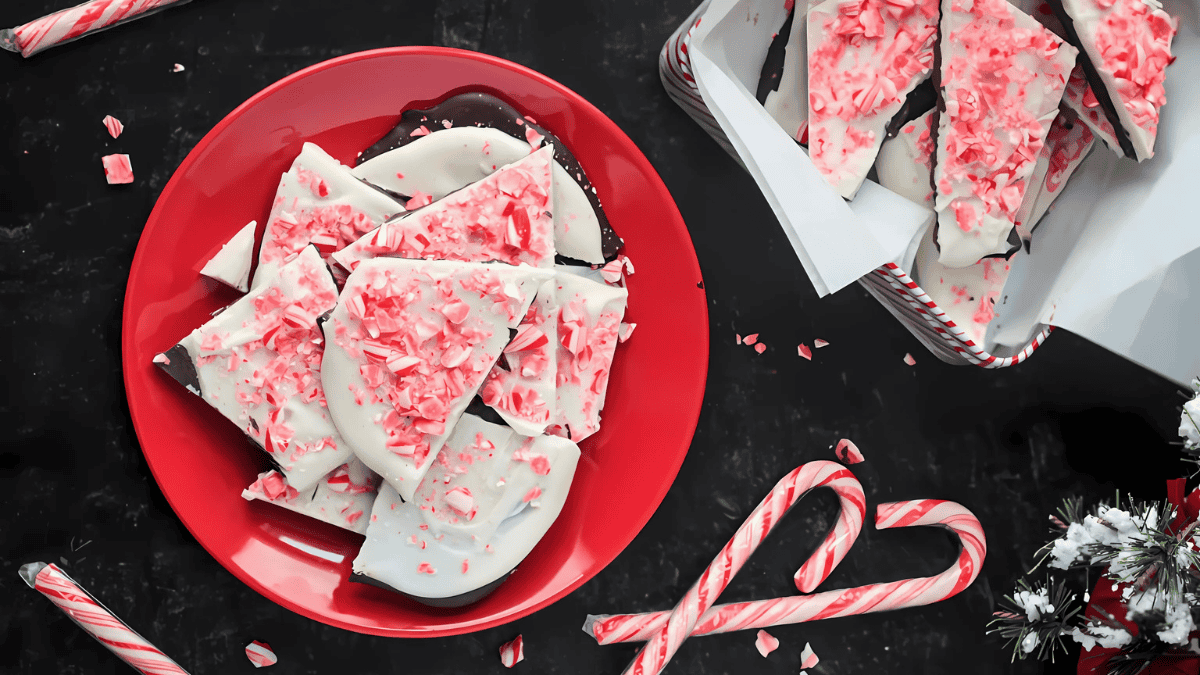 A red plate filled with peppermint bark topped with crushed candy canes sits on a dark table. More peppermint bark is in a nearby white box. Candy canes and peppermint bits are scattered around as decoration.