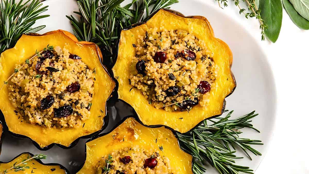 Halved acorn squashes filled with a mixture of quinoa, cranberries, and herbs are arranged on a plate. Fresh rosemary sprigs are placed for garnish.