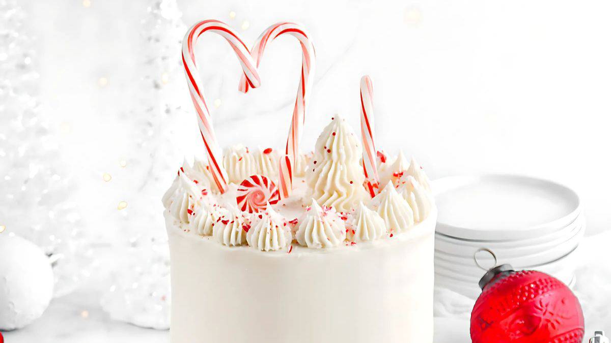 A festive white cake decorated with whipped cream swirls, red sprinkles, and candy canes arranged like a heart on top. In the background, there are white plates and a red ornament, creating a cheerful holiday atmosphere.