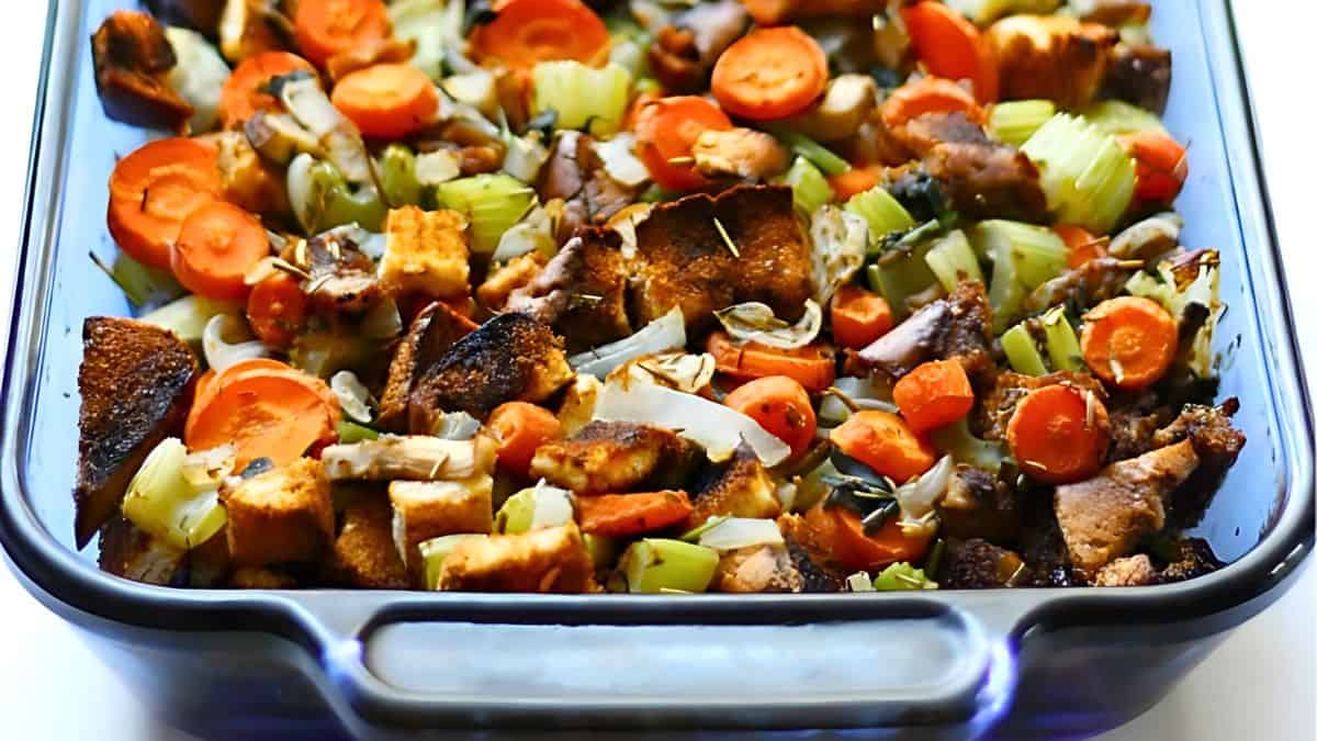 A casserole dish filled with a colorful mixture of roasted vegetables and bread cubes. The dish includes sliced carrots, celery, onions, and crispy bread pieces, all seasoned and baked to a golden brown.