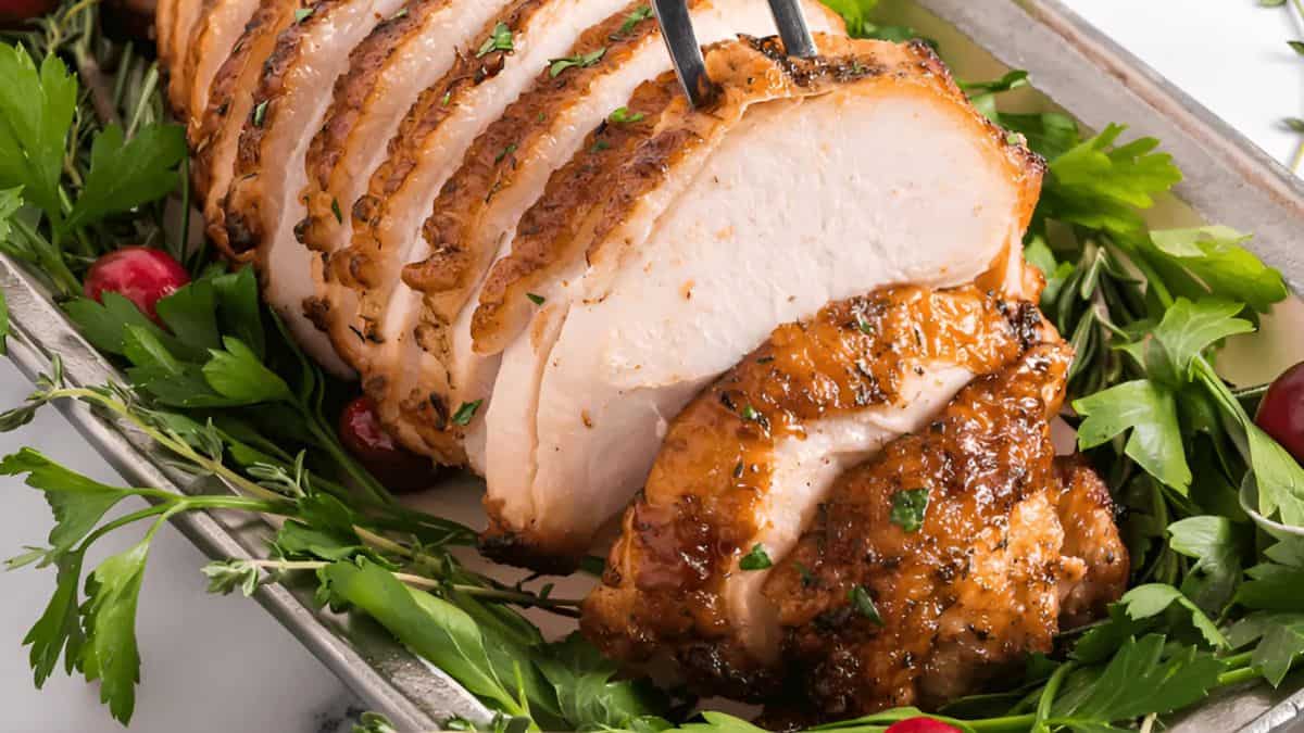 A tray featuring deliciously roasted turkey breast, sliced and garnished with fresh herbs and cranberries. The golden-brown crust showcases a perfectly cooked, juicy interior. A fork is poised over the meat, ready to serve.