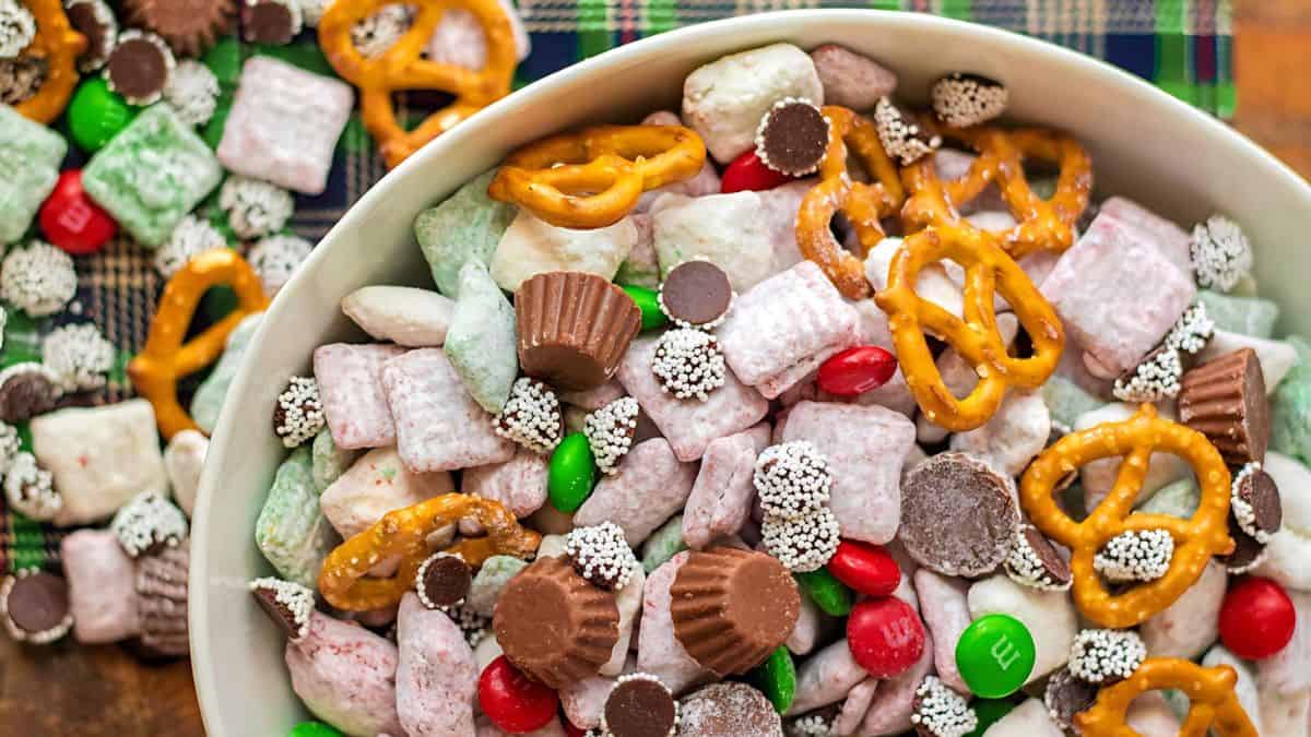 A bowl filled with a mix of colorful snacks, including pretzels, red and green candy-coated chocolates, small peanut butter cups, chocolate-covered candies, and powdered sugar-coated cereal. A few spilled snacks are visible on a checkered cloth.