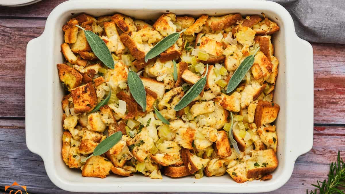 A rectangular dish filled with baked stuffing made of bread cubes, celery, and onions, garnished with sage leaves. The dish sits on a wooden table.
