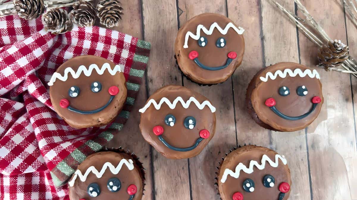 Gingerbread-themed cupcakes with smiling faces, decorated with white zigzag frosting hair, red cheeks, and black eyes, are arranged on a wooden surface. A red and white checkered cloth and small pinecones are nearby.