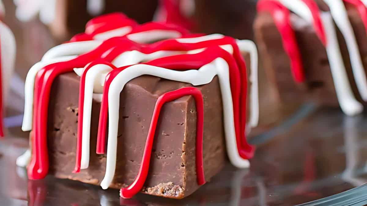 A close-up of a chocolate square treat drizzled with white and red icing, placed on a reflective surface.
