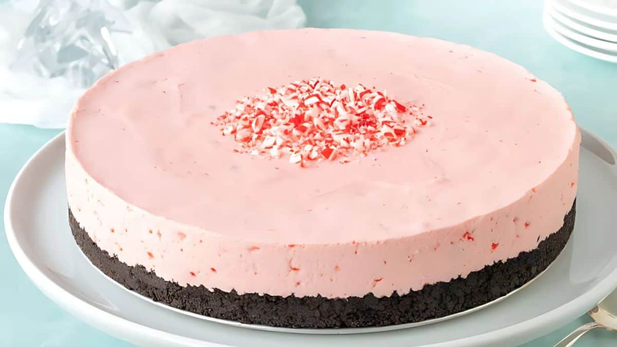 A pink peppermint cheesecake with a chocolate cookie crust sits on a white plate. Crushed peppermint candies are sprinkled in the center. The background features a light blue table and partially visible white cloth and plates.