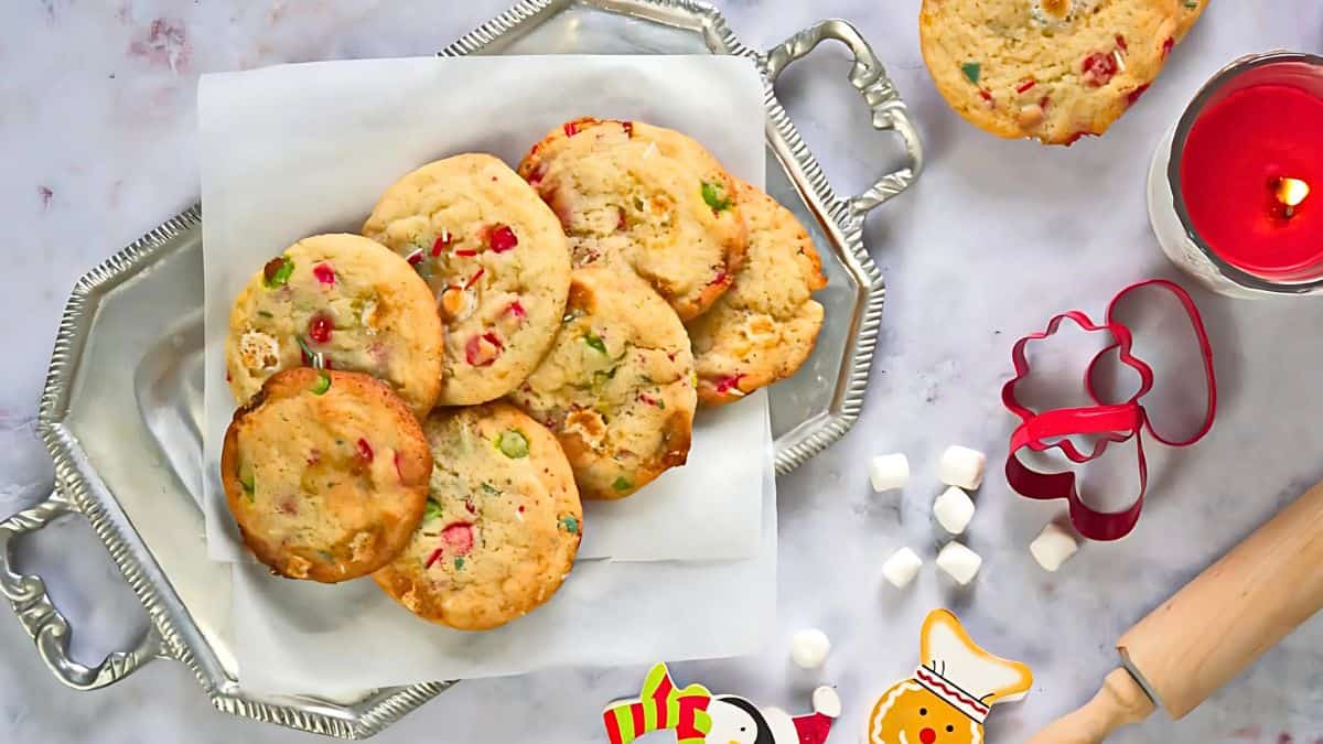 A silver tray with colorful cookies on parchment paper is surrounded by a red candle, gingerbread-themed cookie cutters, mini marshmallows, and a rolling pin on a marble surface. Holiday-themed elements add to the festive ambiance.
