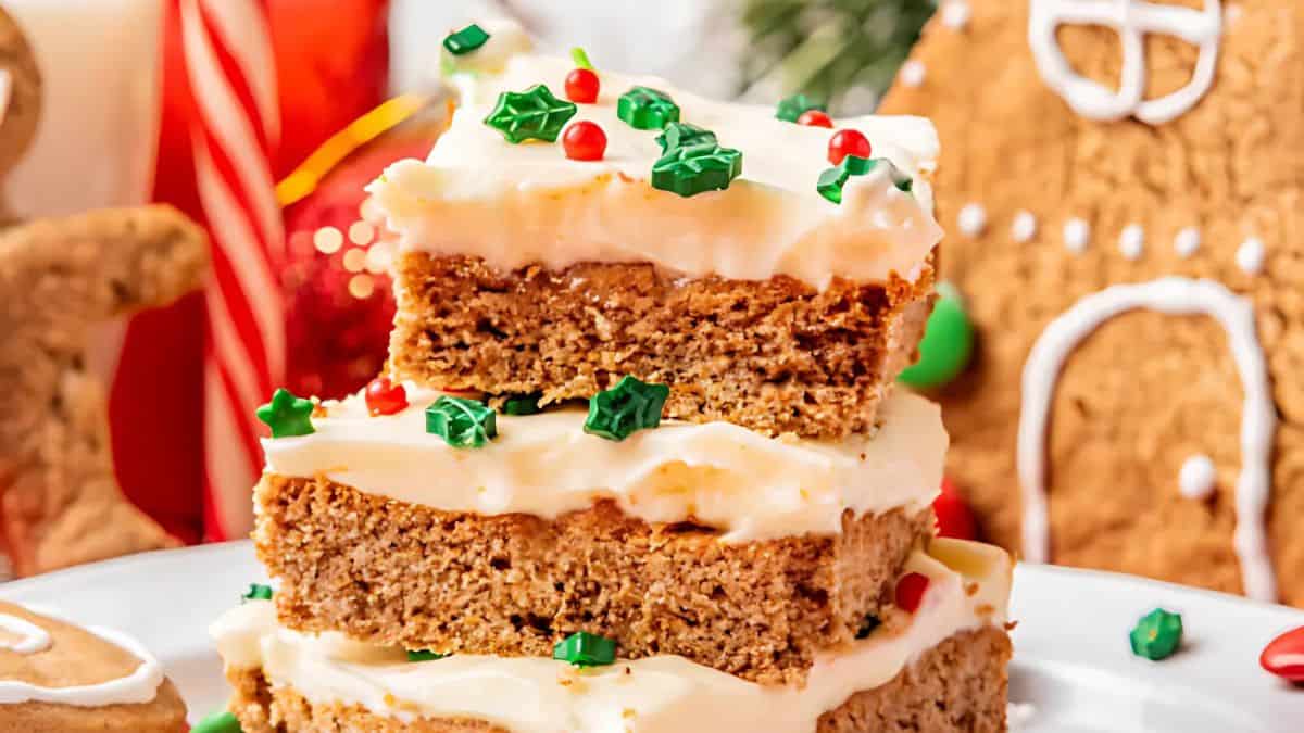 A stack of festive gingerbread bars with creamy frosting, topped with green and red holiday sprinkles. In the background, a gingerbread house and candy cane add to the holiday theme.
