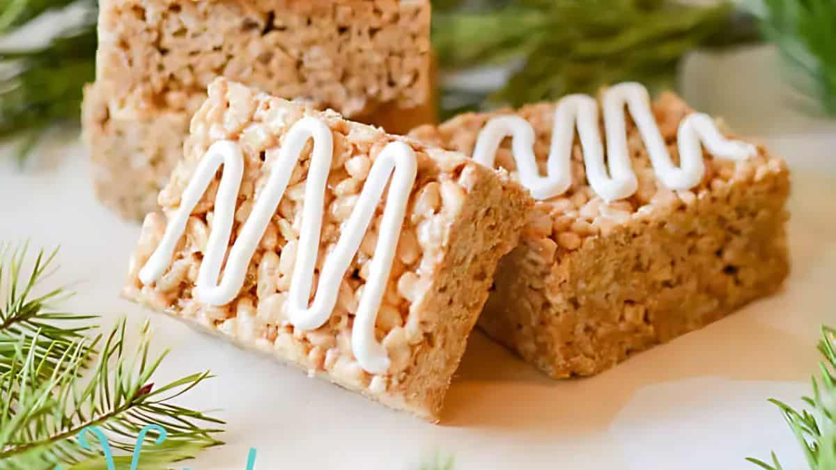 Three rice crispy treats topped with white icing sit on a white surface, surrounded by green pine branches, creating a festive look.
