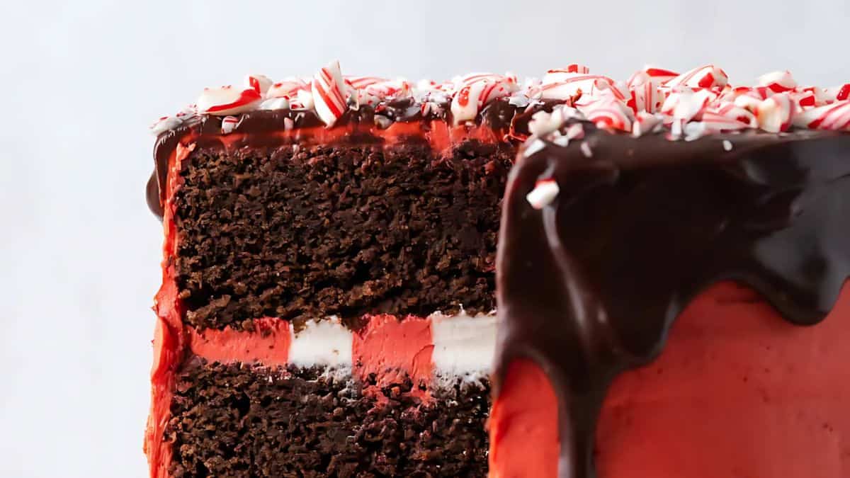 A sliced chocolate peppermint cake with layers of chocolate and red frosting, topped with dark chocolate glaze and sprinkled with crushed peppermint candy.
