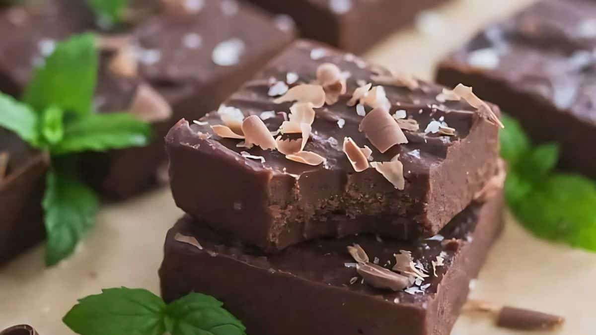 Close-up of two stacked chocolate fudge squares topped with chocolate shavings. The top square has a bite taken out of it. Fresh mint leaves are scattered around on a light-colored surface.