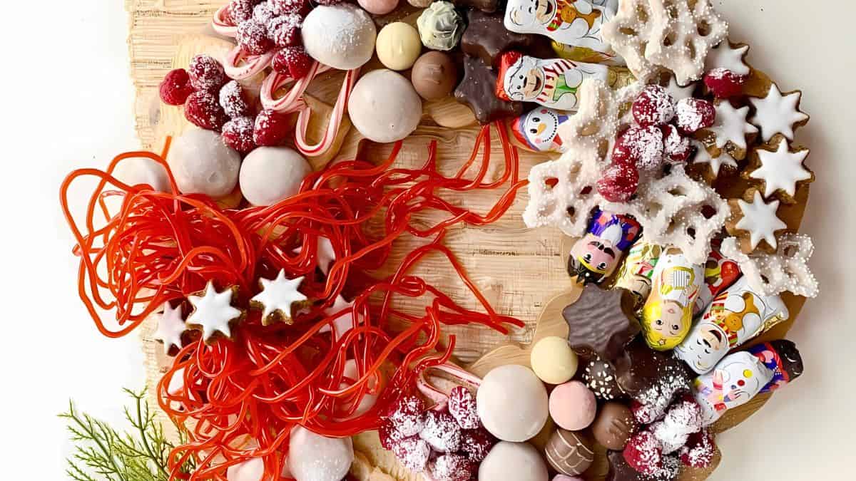 A festive wreath made of assorted sweets and treats, including colorful candy canes, chocolates wrapped in festive foil, white cookies, red licorice, and chocolate stars, arranged on a wooden surface. A small sprig of greenery is visible on the side.