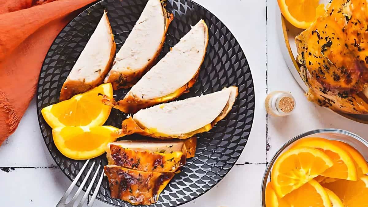 A black plate with sliced roasted turkey breast and three wedges of orange. A small glass bowl with more orange slices is nearby, and an orange napkin is partially visible in the background.