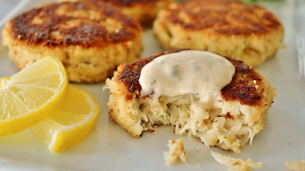 Close-up of crab cakes on a white plate, one with a bite taken out, revealing tender crab meat. Topped with creamy sauce, accompanied by lemon wedges for garnish. Golden brown crust suggests a crispy texture.