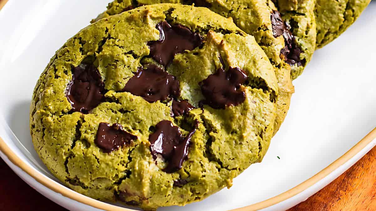 Close-up of matcha green tea chocolate chip cookies on a white plate with a wooden rim. The cookies are green and studded with dark chocolate chunks, creating a contrasting visual appeal.