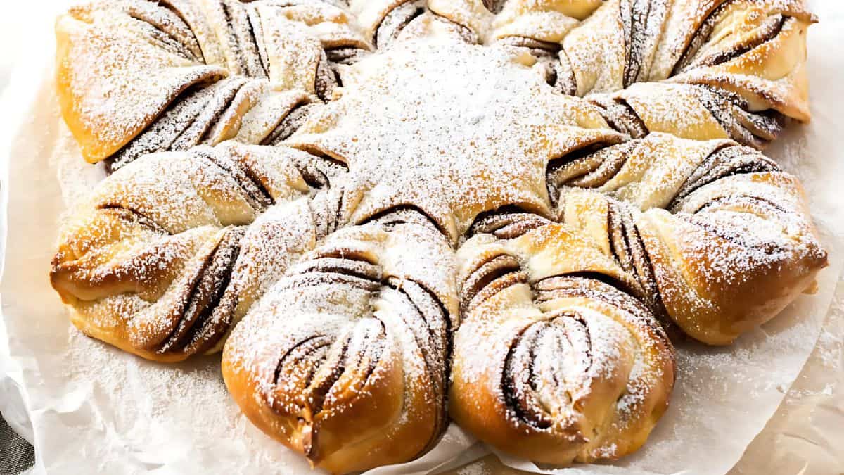 A star-shaped, braided pastry dusted with powdered sugar. The pastry is golden brown, with visible layers of chocolate filling in the twists. It sits on a piece of parchment paper.