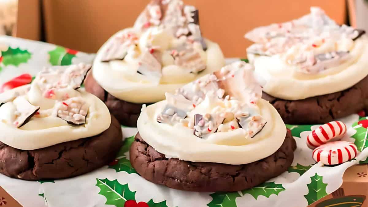 Four chocolate cookies topped with white frosting and peppermint candy pieces on holiday-themed paper, alongside striped peppermint candies.