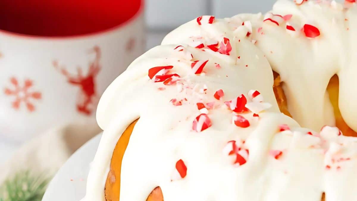 A festive bundt cake topped with creamy white icing and sprinkled with crushed red and white peppermint candy pieces, with a holiday-themed mug in the background.