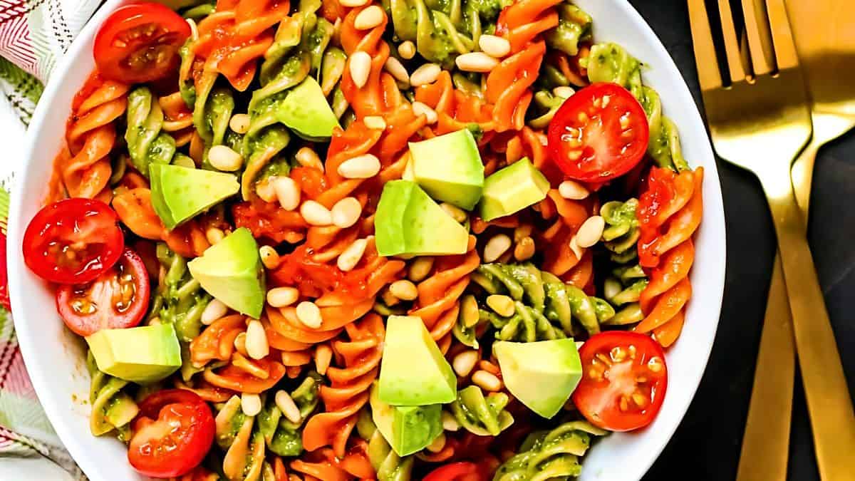 A colorful bowl of pasta salad featuring tri-color rotini, cherry tomatoes, avocado chunks, and pine nuts. Gold utensils are placed beside the bowl.