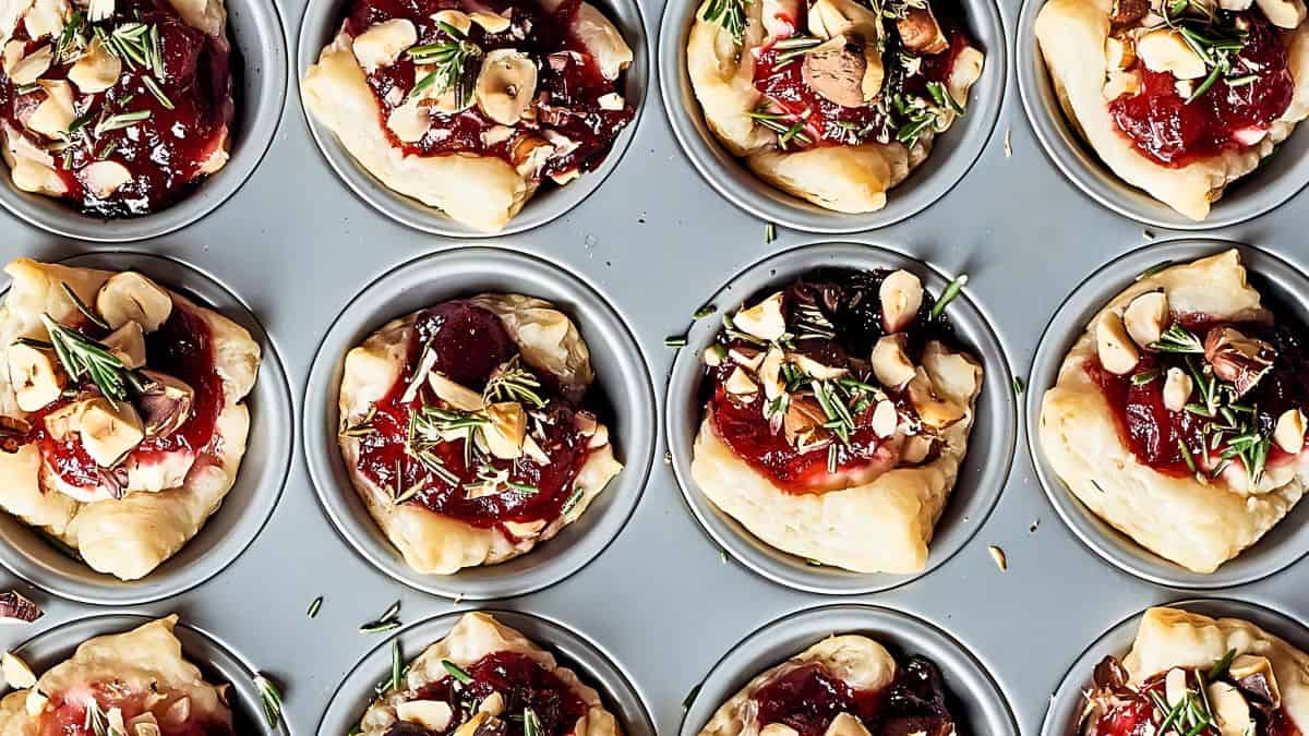 Close-up of a muffin tray filled with baked appetizers. Each portion consists of puff pastry topped with a red jam, chopped nuts, and fresh herbs, arranged in a neat grid.