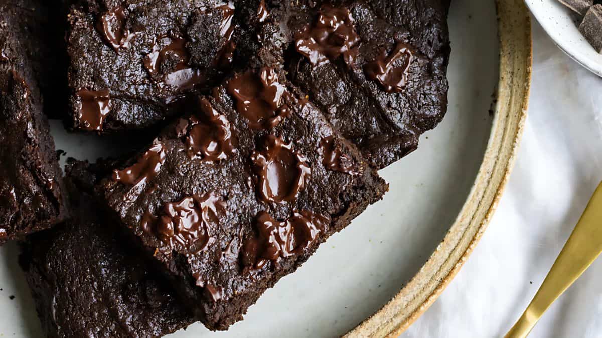Close-up of rich, dark chocolate brownies on a plate. The brownies have a glossy finish with melted chocolate chunks visible on the surface, creating a decadent and indulgent appearance. A fork is partially visible next to the plate.