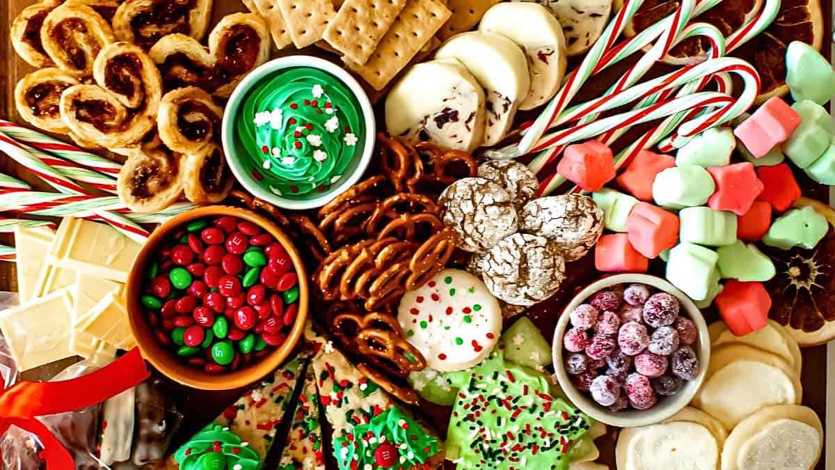 A festive assortment of holiday treats, including candy canes, green frosted cookies, pretzels, chocolate crinkle cookies, red and green candies, marshmallows, and frosted cookies on a platter.