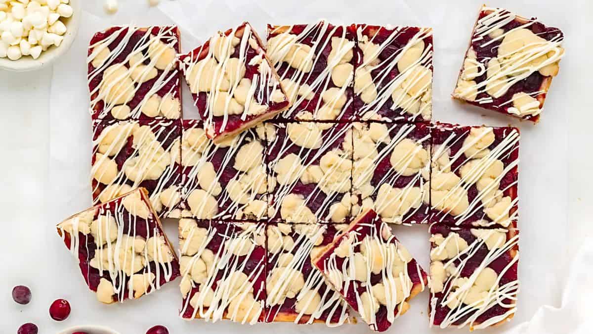 A batch of cranberry almond bars topped with white chocolate drizzle. The bars are square-shaped and placed on parchment paper. Some almonds and cranberries surround the bars, enhancing the presentation.