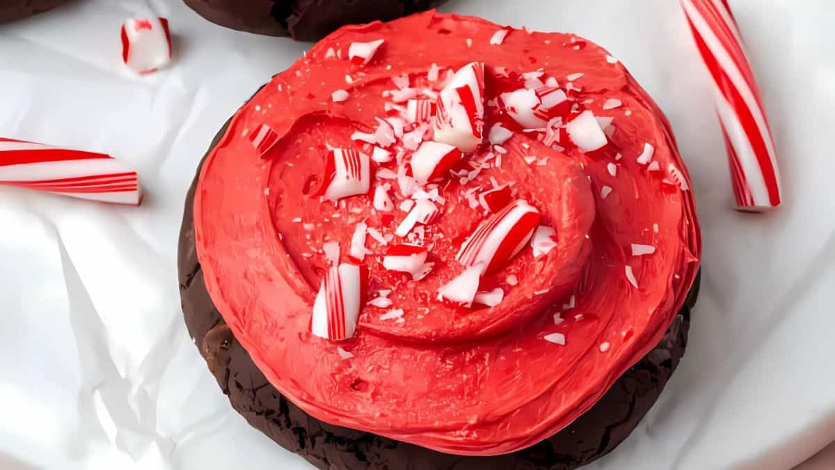 A chocolate cookie topped with red frosting and sprinkled with crushed peppermint candy pieces. The cookie is on a sheet of white parchment paper, surrounded by broken pieces of red and white candy canes.