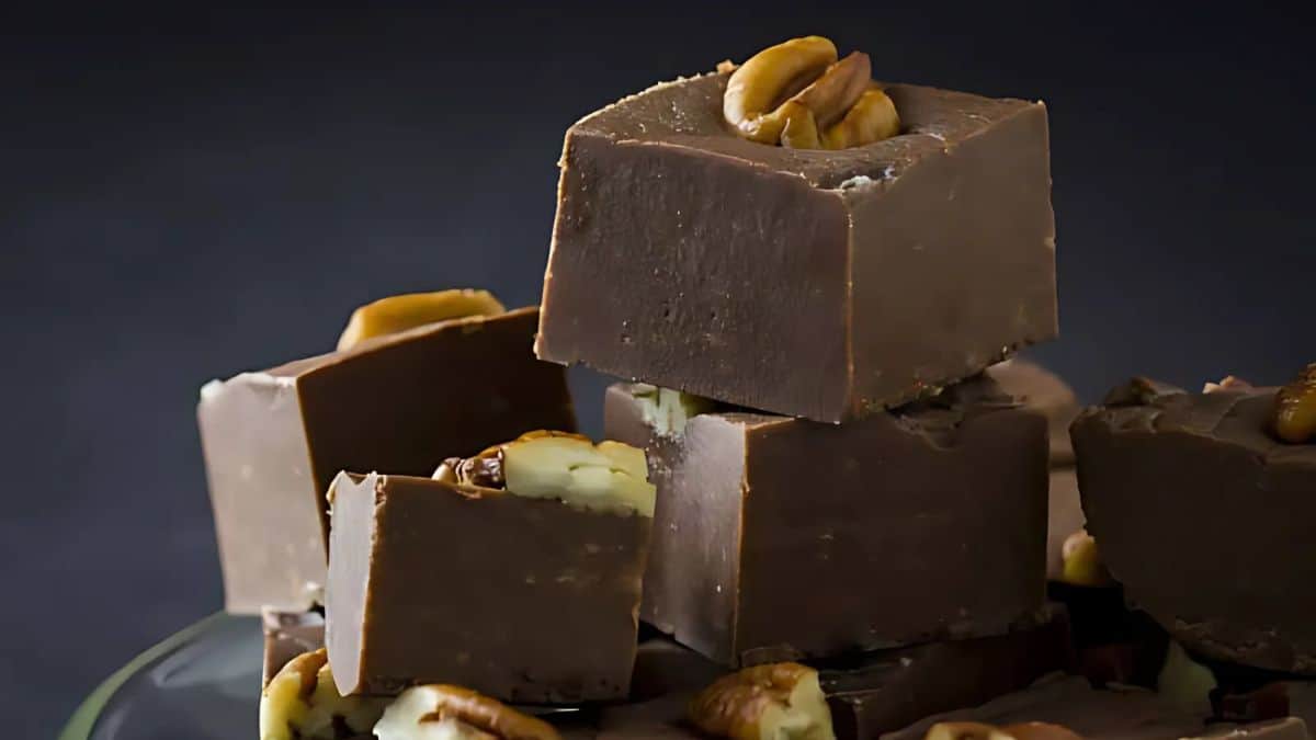 A stack of rich, creamy chocolate fudge pieces topped with pecans. The dark background contrasts with the smooth texture of the fudge, highlighting its decadent appearance.