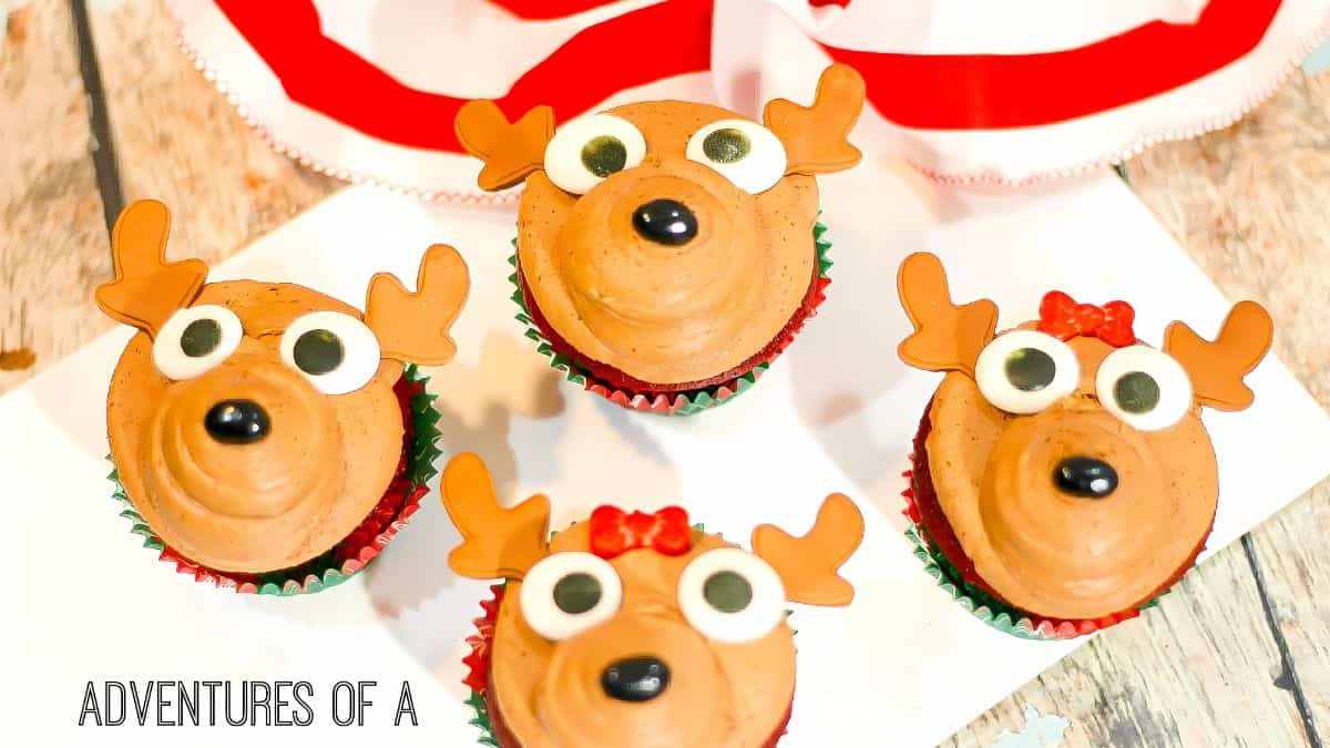 Four cupcakes decorated as reindeer with brown frosting, candy eyes, red noses, and pretzel antlers are arranged on a wooden surface next to a red and white striped cloth. One reindeer has a red bow.