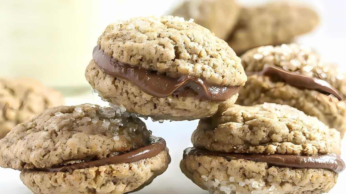 Close-up of oatmeal cookie sandwiches filled with chocolate cream. The cookies have a golden-brown texture and a sprinkle of sugar crystals on top. Several cookies are stacked, displaying the creamy filling.