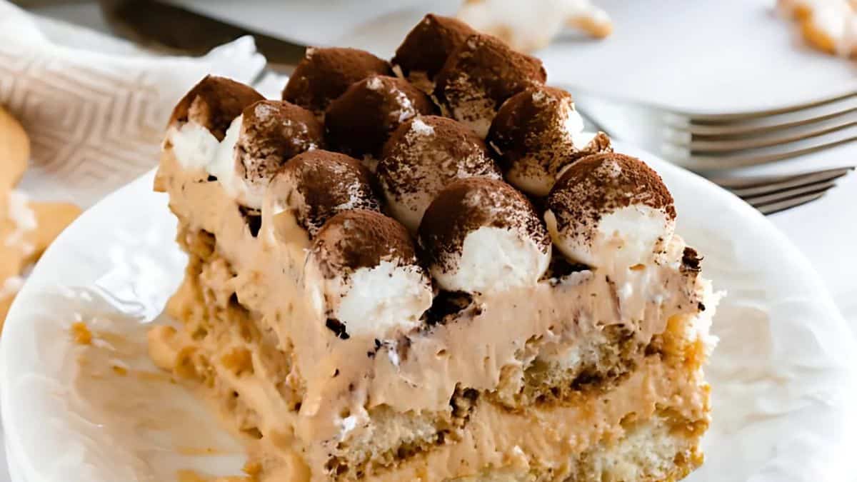 A close-up of a slice of tiramisu on a white plate. The dessert features layers of coffee-soaked ladyfingers and creamy mascarpone, topped with cocoa powder and dollops of whipped cream. Forks are visible in the background.