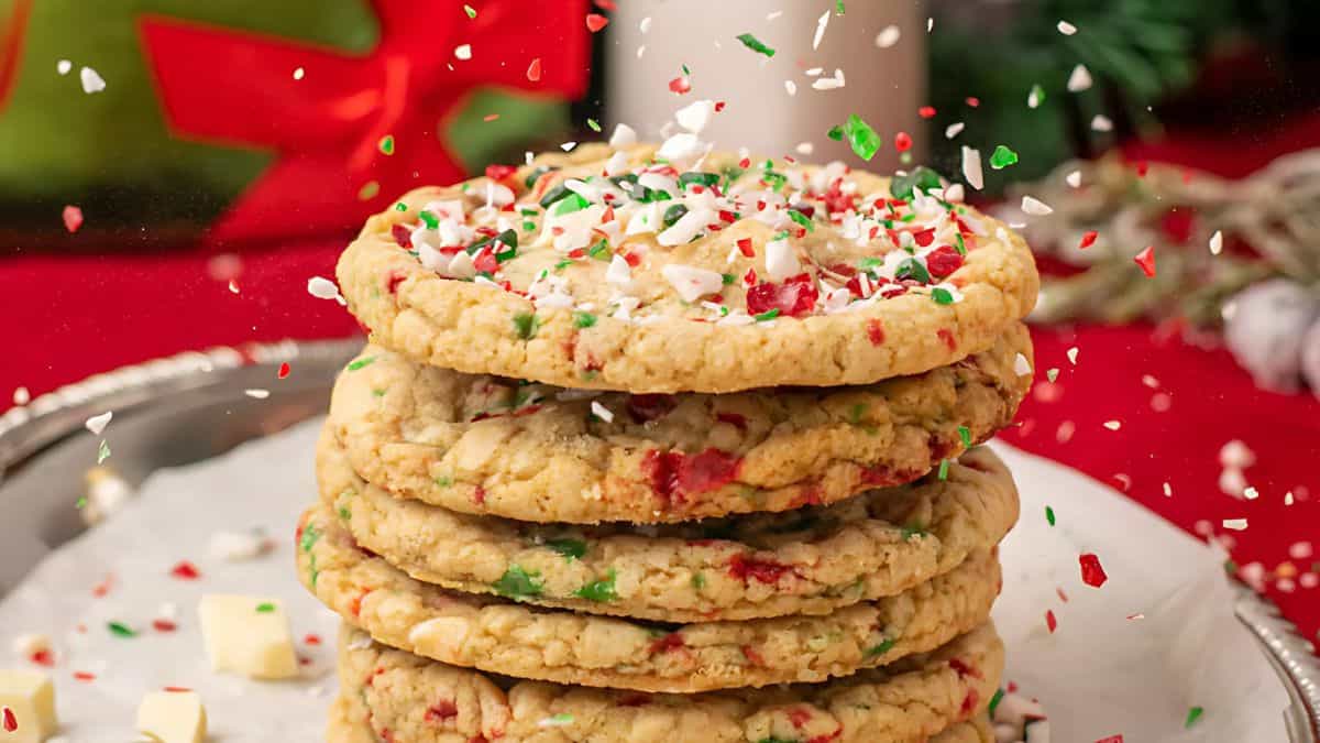 A stack of festive cookies topped with colorful sprinkles sits on a plate. The cookies contain visible red and green pieces, and scattered sprinkles add a festive touch. A blurred holiday-themed background enhances the seasonal atmosphere.