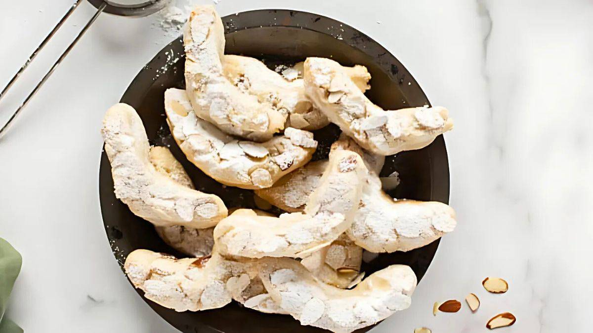 A plate of crescent-shaped cookies dusted with powdered sugar, garnished with sliced almonds, on a marble surface. A metal sieve is partially visible at the top.