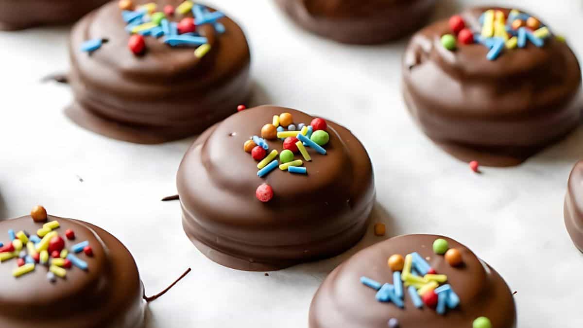 Chocolate-covered cookies topped with colorful sprinkles are arranged on a light surface. These treats have a smooth, glossy finish, and the vibrant sprinkles add a playful touch to each cookie.