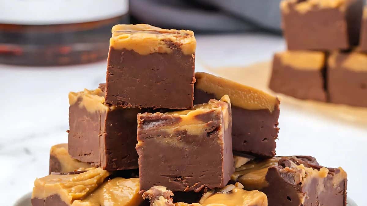 A stack of chocolate and peanut butter fudge squares on a white surface, with more fudge pieces in the background. The fudge has a rich chocolate base topped with a swirl of creamy peanut butter.